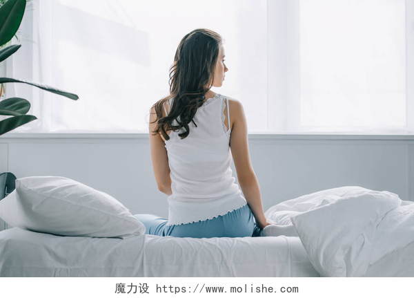 一个女人坐在白色的床铺上坐在床上看着的穿着睡衣的年轻女子的背部视图 